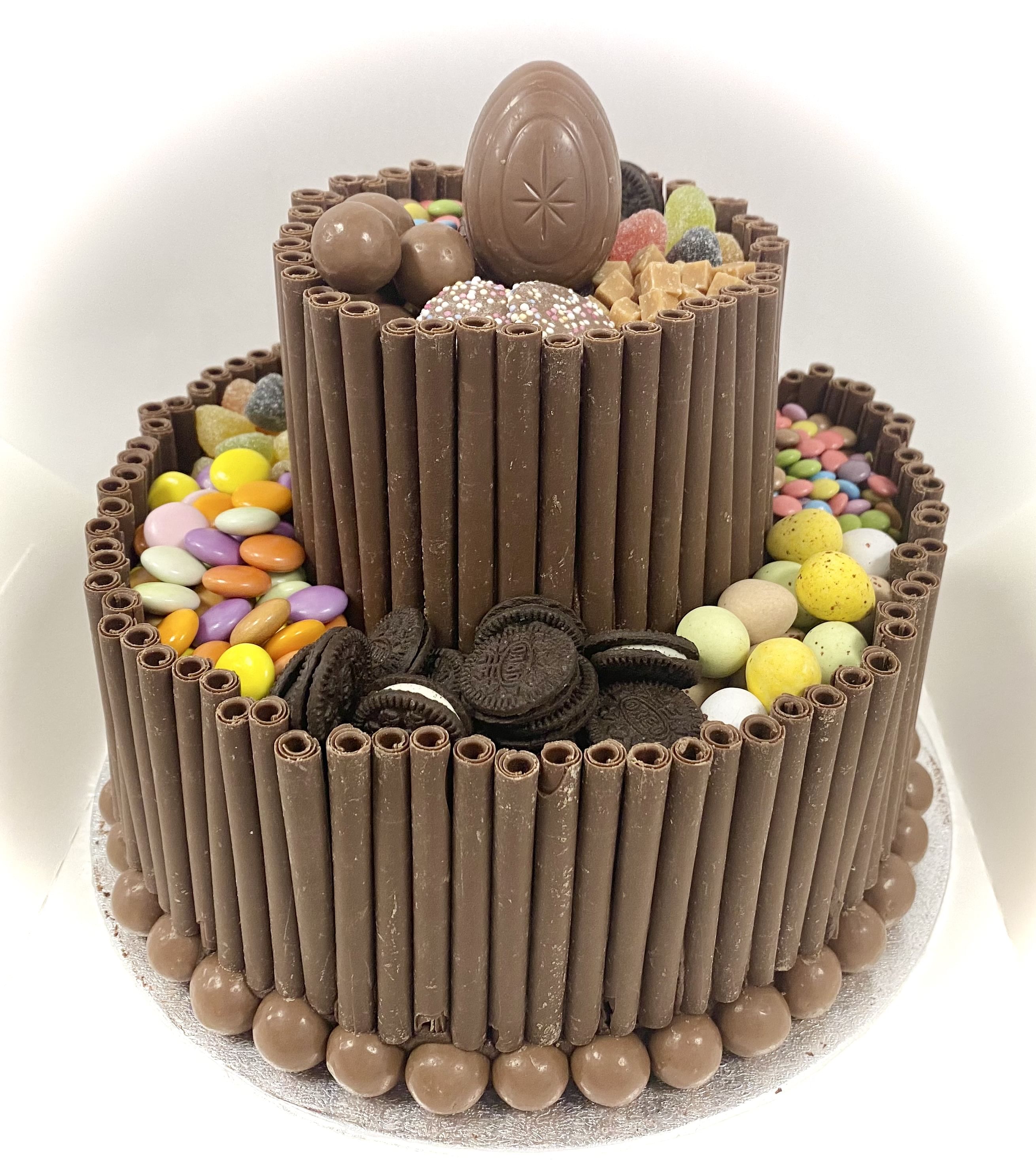 A two-tier Chocolate Blackout Cake adorned with chocolate sticks around the edges. The cake is topped with a large chocolate egg and decorated with an assortment of candies, including colorful sugar-coated chocolates, jelly candies, and mini chocolate eggs, all held together by luscious chocolate frosting.